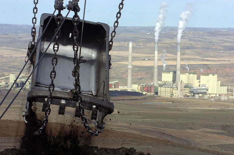 Image of a large metallic shovel suspended from chains in the foreground, with a coal generating plant in the background.
