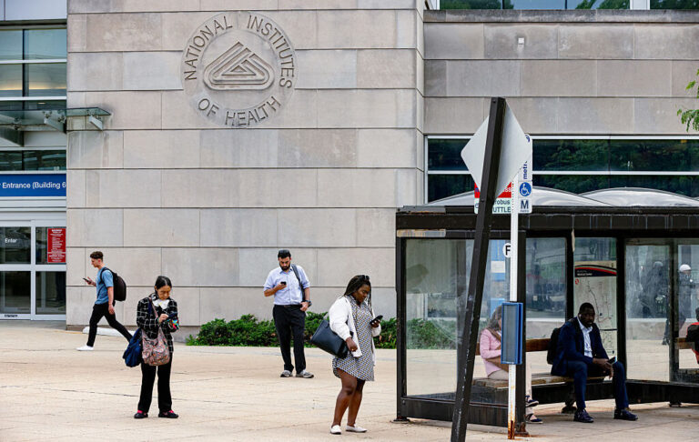 People standing near a bus stop in front of a large stone building facade with the NIH logo etched into the stone.