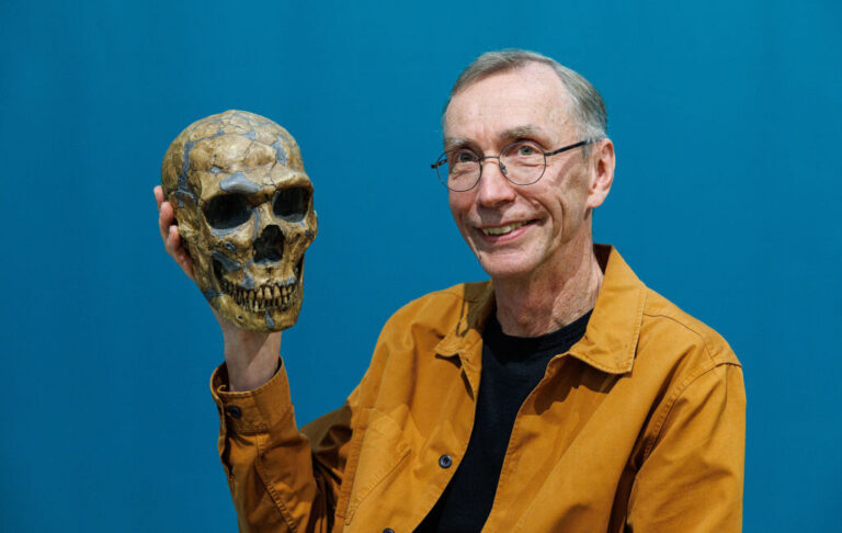 Image of a man with glasses and a mustard colored jacket holding up a skull.